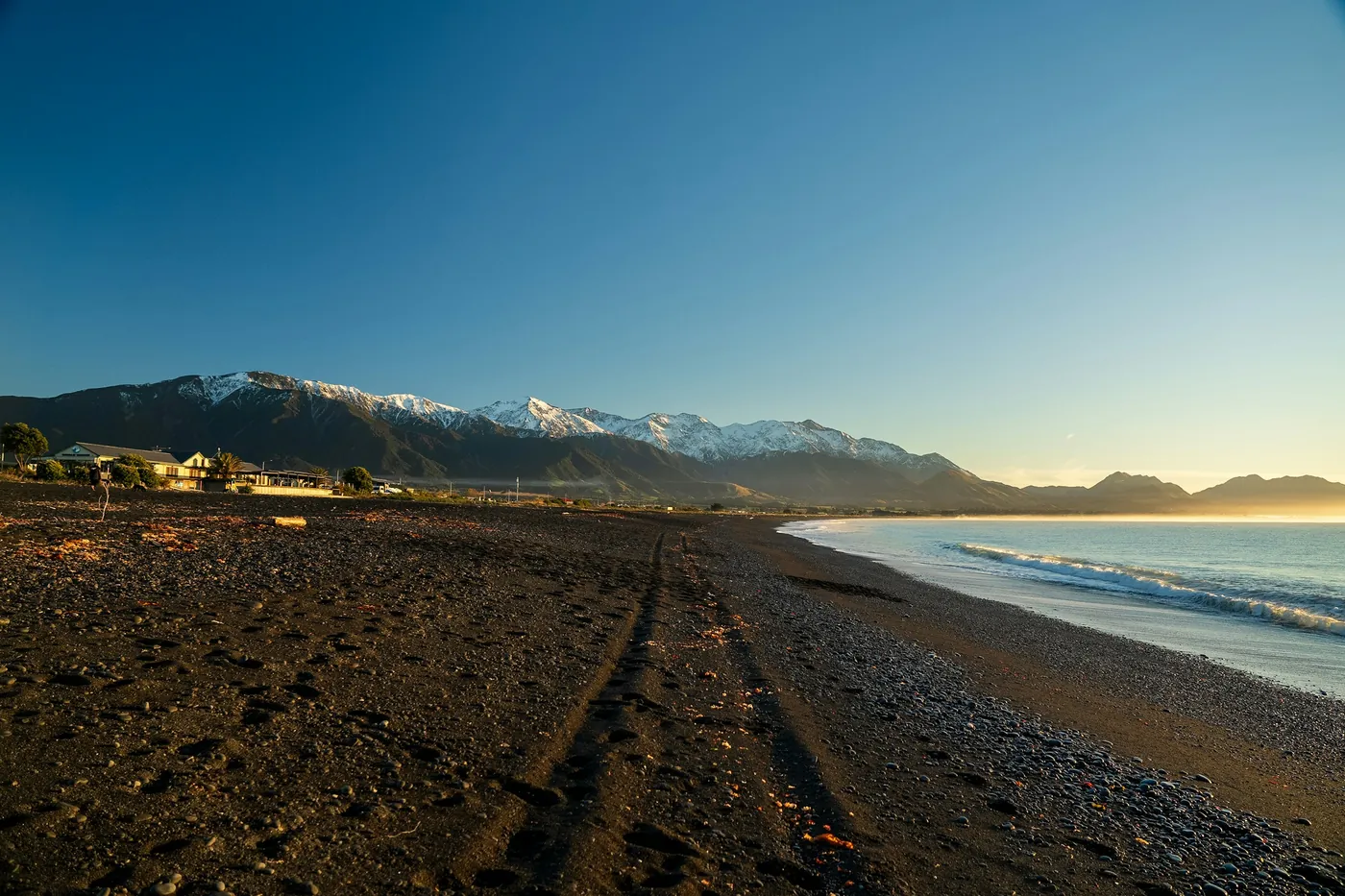 A wide, dark pebble beach with tyre tracks leading towards the sea, low houses and snow-dusted mountains in warm evening light
