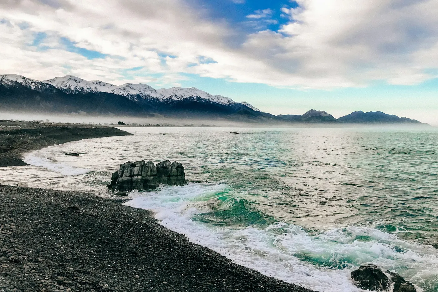 Waves breaking on a grey pebble shore with a rock outcrop in the water and a snow-covered mountain range along the coast