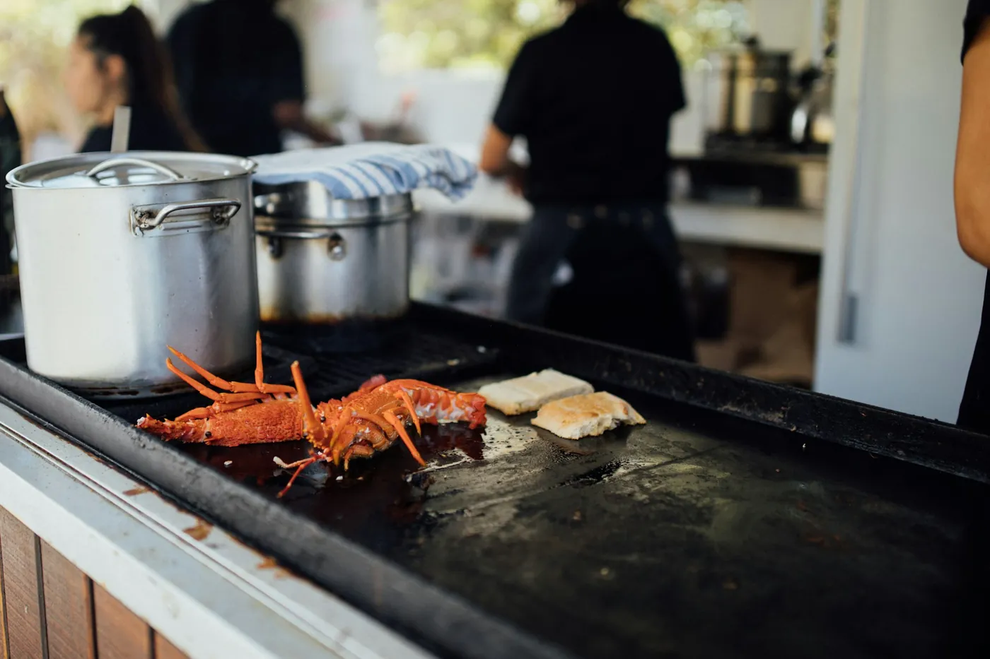 A red spiny lobster cooking on a flat grill at a food stall next to steel pots, with staff blurred in the background