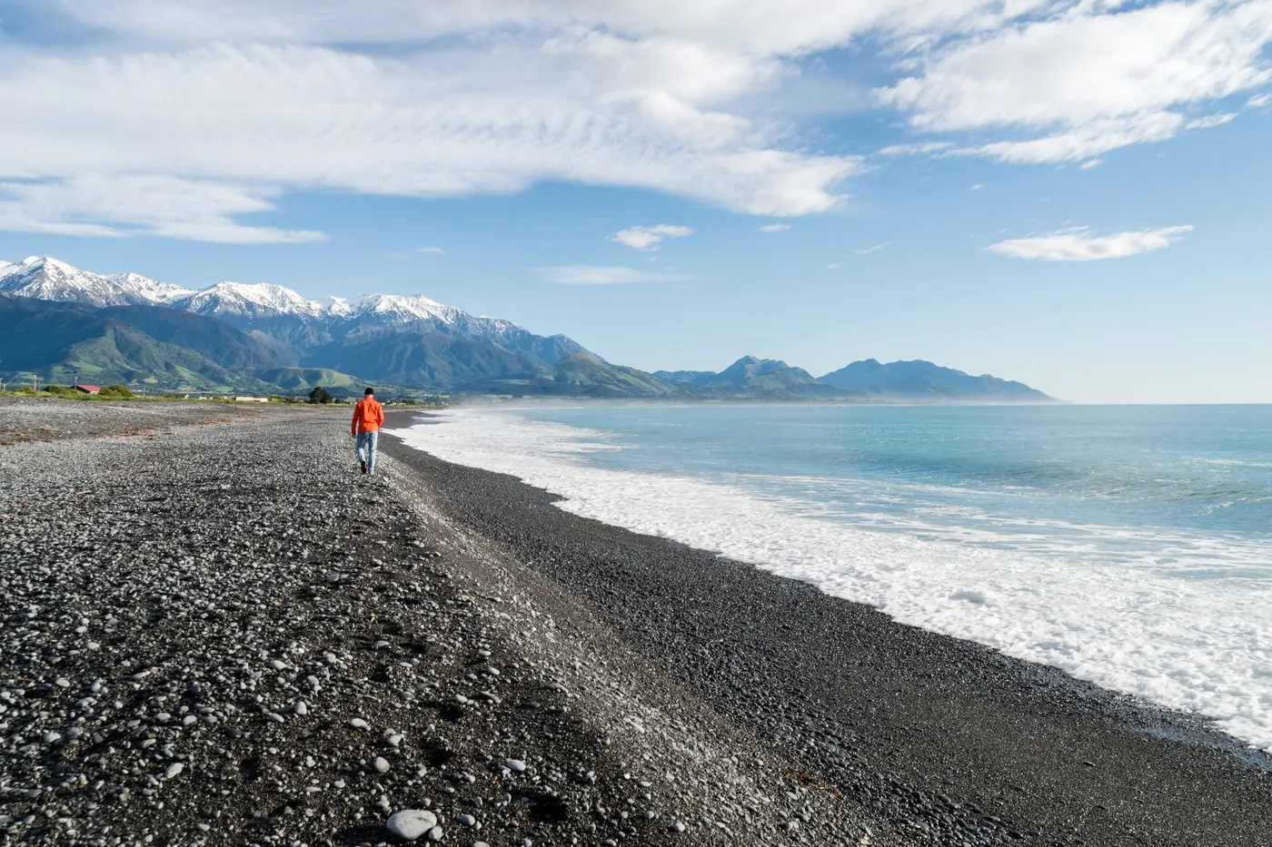 A person walking along a dark pebble beach beside white surf, with snow-capped mountains on the horizon under a blue sky with clouds
