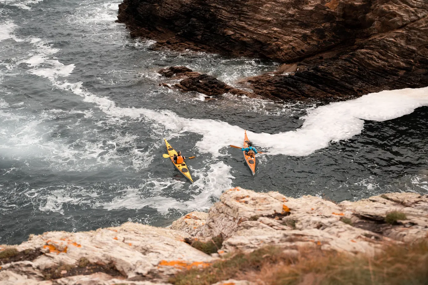 Two sea kayakers paddling through white water between dark rocks and a rocky ledge on a sunny day