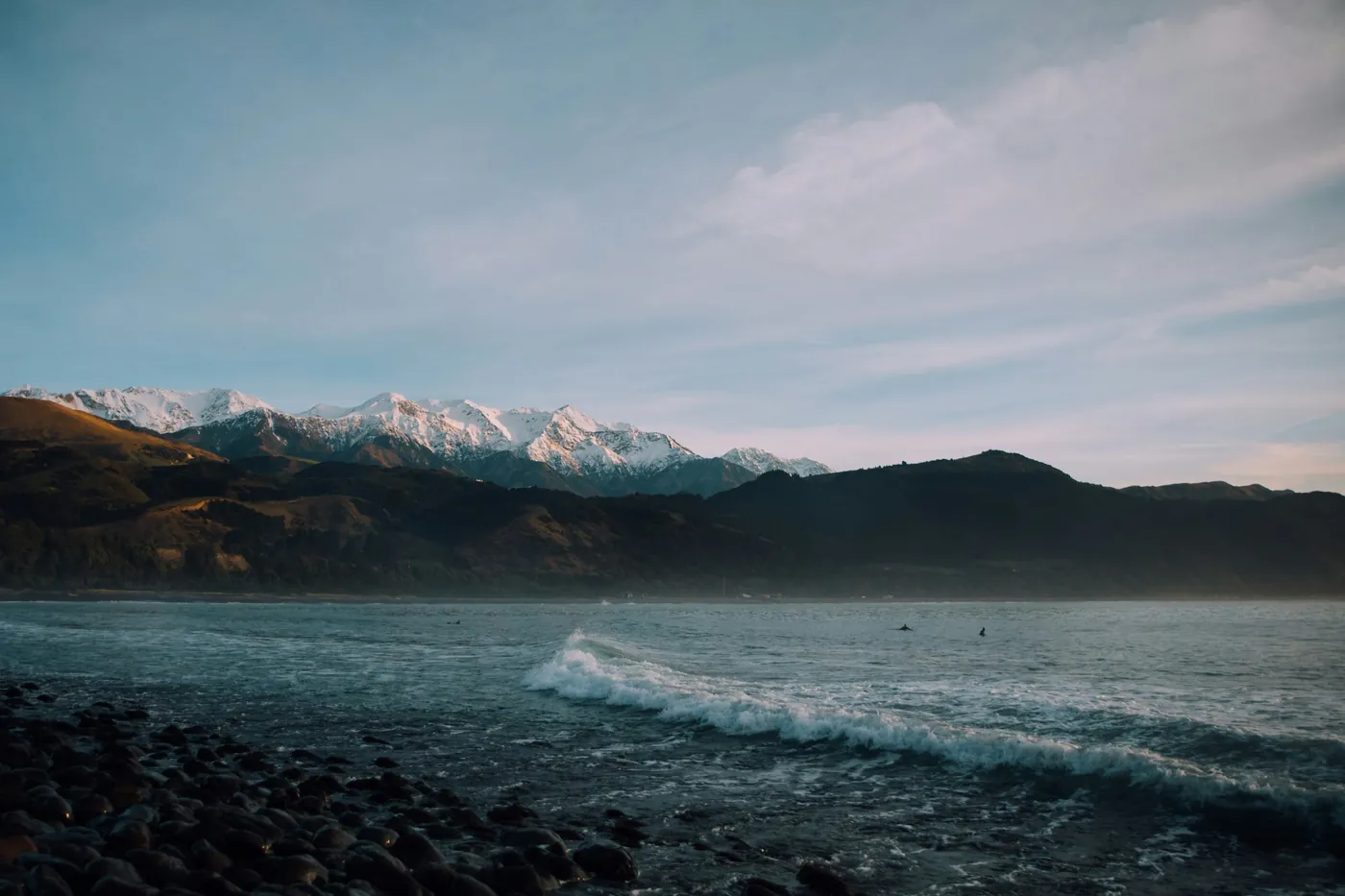 Snow-capped mountains behind dark foothills, with a wave rolling in over a stony beach in the foreground