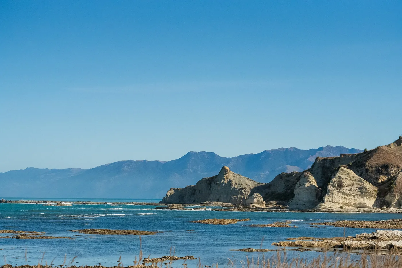 Pale limestone cliffs and a flat rock platform at the edge of a blue sea, with a mountain range in the distance