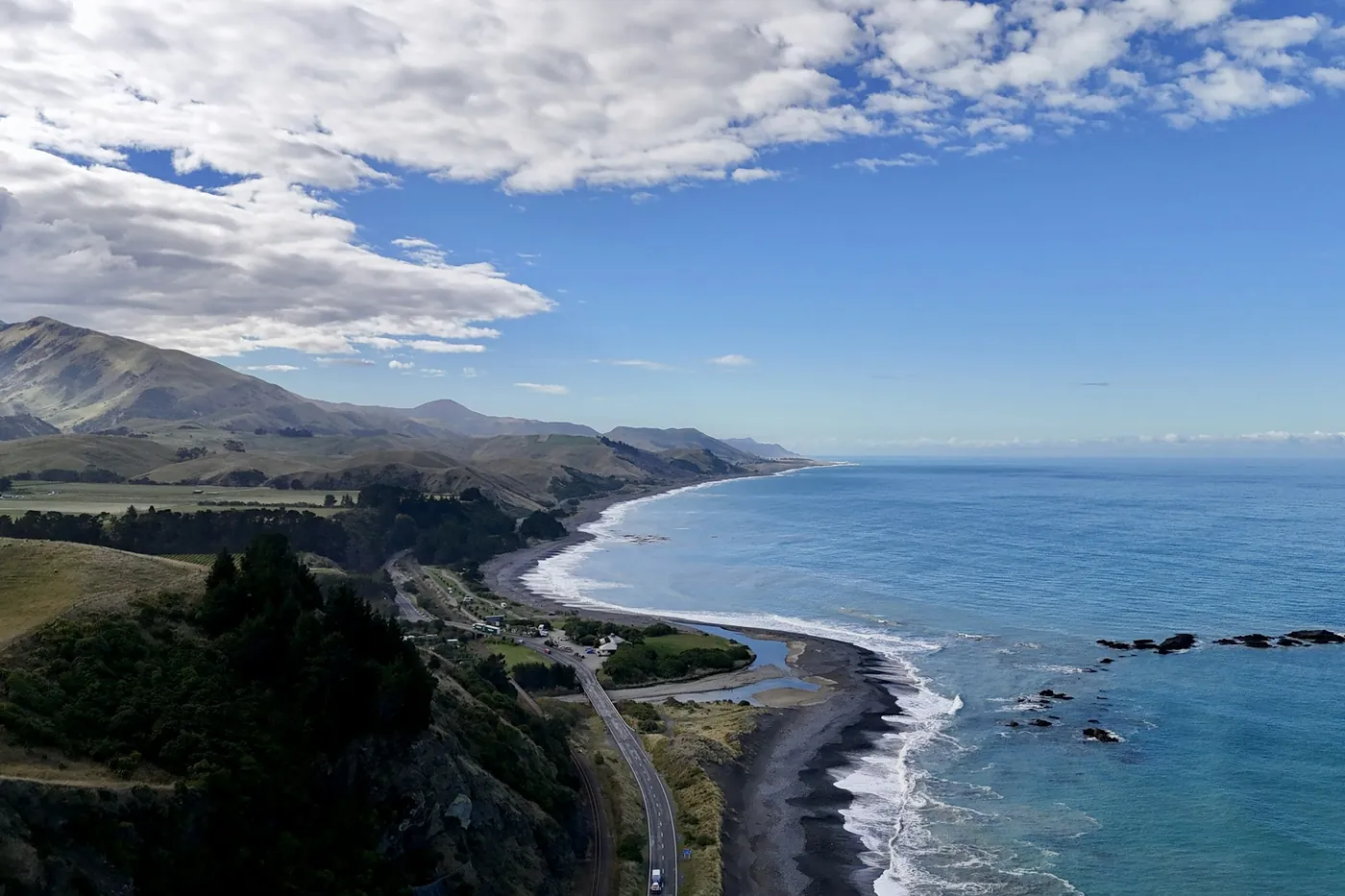 Aerial view of a coastal highway and railway line running between green hills and a dark sand beach, with the sea stretching to the horizon