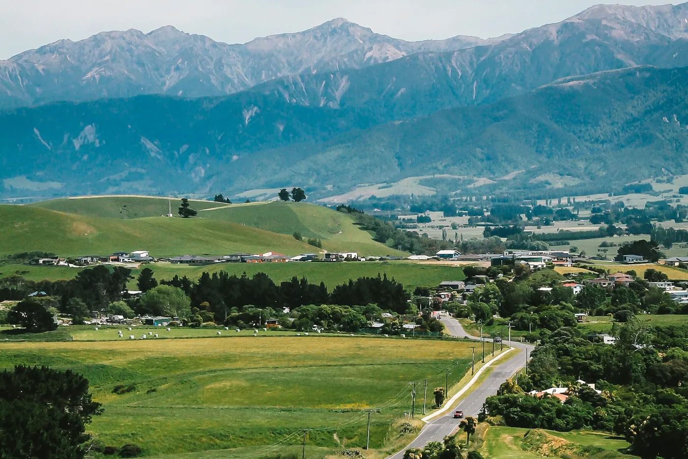Green farmland and the houses on the edge of a small town on a plain, below a mountain range, with a winding road in the foreground